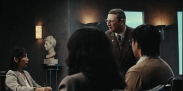 A man in a suit stands and speaks to three seated people in a dimly lit office, with a bust sculpture in the background, evoking the quirky dynamics of a workplace comedy starring Tim Robinson.