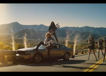 Two people with musical instruments stand on a silver car parked on a mountain road, sparks flying behind them like a Bon Jovi concert, while six women pose in the background—an epic scene worthy of a State Farm Super Bowl 2026 ad.