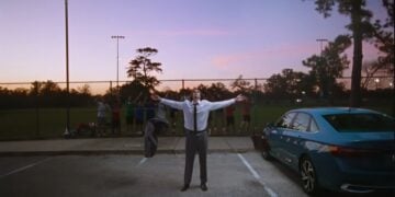 A man stands in a parking lot with arms outstretched, tossing his jacket, as nostalgia fills the air. Behind a fence, people watch near a blue car at sunset—capturing a moment reminiscent of classic Volkswagen Super Bowl 2026 spirit.