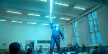A person in a blue jumpsuit stands on a table in a cafeteria, reaching up as a bright blue beam of light shoots down onto their raised hand, while others watch—a striking scene inspired by the DHSC Smokers’ Campaign.